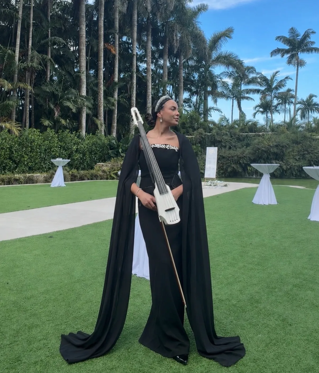 Saydee Sarduy in a black dress holding her electric cello in a palm garden.